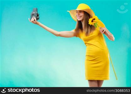 summer woman wearing yellow dress and hat with sunflower taking self picture with old vintage camera on vivid blue background