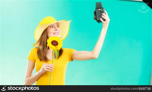 Summer woman wearing yellow dress and hat with sunflower taking self picture with old vintage camera on vivid blue background