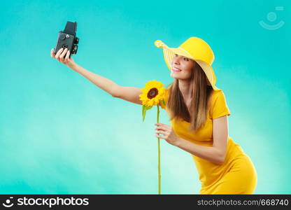 summer woman wearing yellow dress and hat with sunflower taking self picture with old vintage camera on vivid blue background