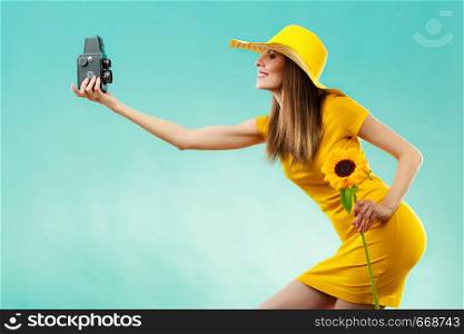 summer woman wearing yellow dress and hat with sunflower taking self picture with old vintage camera on vivid blue background