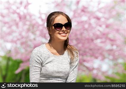 summertime, leisure and people concept - happy young woman in sunglasses in garden over cherry blossom background. woman in sunglasses in garden over cherry blossoms