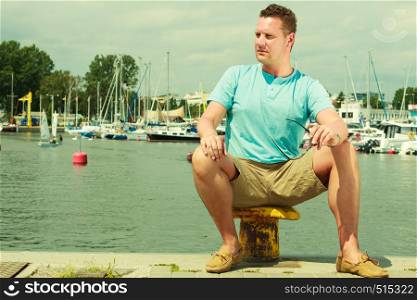 Summertime vacation adventure concept. Man spending his free time walking on marina, sightseeing during summer, guy sitting on bitt.. Man walking on marina during summer