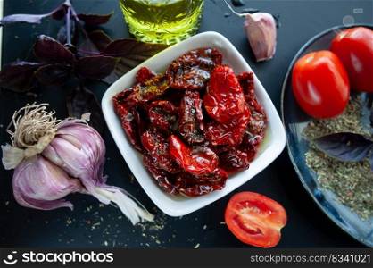 Sun-dried tomatoes in a bowl surrounded by ingredients, spices , garlic and basil to prepare. Rustic style, top view  