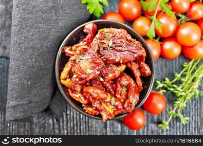 Sun-dried tomatoes in oil with thyme and basil in a bowl, napkin, fresh small tomatoes and parsley on wooden board background from above