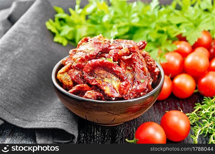 Sun-dried tomatoes in oil with thyme and basil in a bowl, napkin, fresh small tomatoes and parsley on dark wooden board background