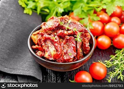 Sun-dried tomatoes in oil with thyme and basil in a bowl, napkin, fresh small tomatoes and parsley on wooden board background