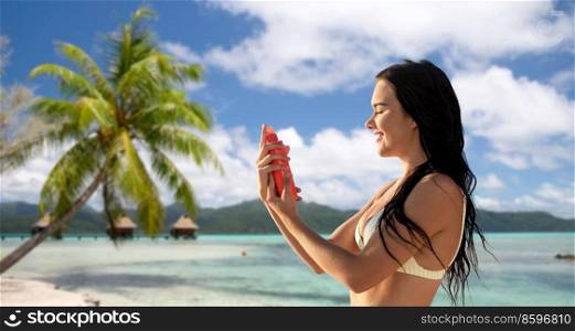 sun protection, tanning and beauty concept - happy smiling young woman in bikini swimsuit spraying sunscreen spray on her face over tropical beach background in french polynesia. smiling woman in bikini with sunscreen on beach