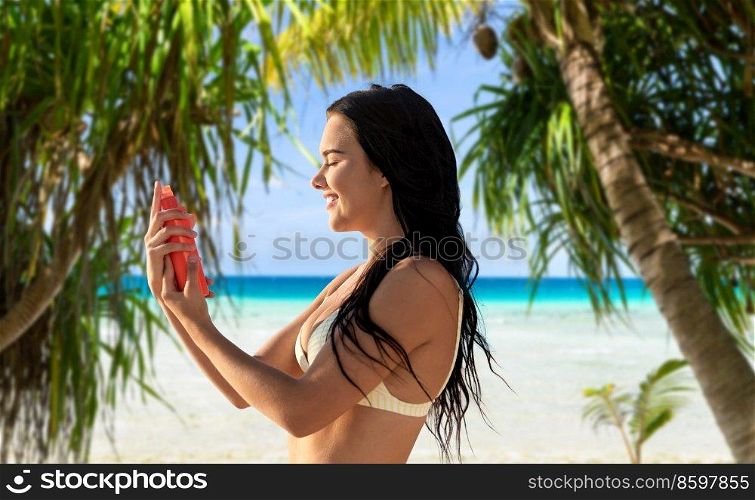 sun protection, tanning and beauty concept - happy smiling young woman in bikini swimsuit spraying sunscreen spray on her face over tropical beach and palm trees background in french polynesia. smiling woman in bikini with sunscreen on beach