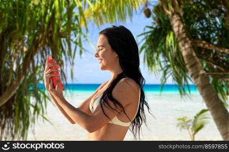sun protection, tanning and beauty concept - happy smiling young woman in bikini swimsuit spraying sunscreen spray on her face over tropical beach and palm trees background in french polynesia. smiling woman in bikini with sunscreen on beach
