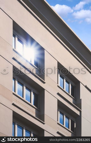 Sunlight reflection on surface of multiple blue glass windows on gray office building wall against white cloud and blue sky in vertical frame