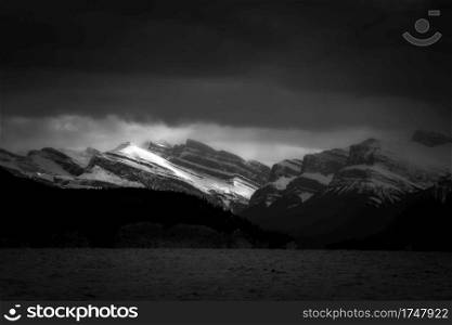 Sunlight streams through the dark clouds illuminating the snow capped mountains in the distance across Abraham Lake in Alberta, Canada