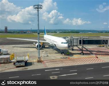 Sunny day at the Indonesian airport. An aircraft with an attached jet bridge for boarding and disembarking passengers. Ocean and jungle in the background. Airplane in Indonesian Airport With Jet Bridge for Passengers