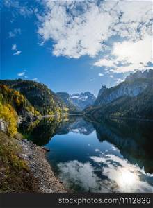 Sunny idyllic colorful autumn alpine view. Peaceful mountain lake with clear transparent water and reflections. Gosauseen or Vorderer Gosausee lake, Upper Austria. Dachstein summit and glacier in far.