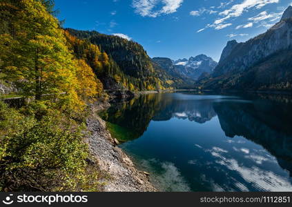 Sunny idyllic colorful autumn alpine view. Peaceful mountain lake with clear transparent water and reflections. Gosauseen or Vorderer Gosausee lake, Upper Austria. Dachstein summit and glacier in far.