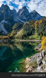 Sunny idyllic colorful autumn alpine view. Peaceful mountain lake with clear transparent water and reflections. Gosauseen or Vorderer Gosausee lake, Upper Austria. Dachstein summit and glacier in far.