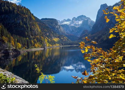 Sunny idyllic colorful autumn alpine view. Peaceful mountain lake with clear transparent water and reflections. Gosauseen or Vorderer Gosausee lake, Upper Austria. Dachstein summit and glacier in far.