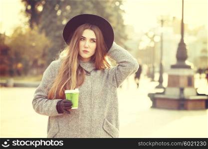 Sunny lifestyle portrait of young stylish hipster woman walking on street, wearing cute trendy hat, drinking hot latte coffee outdoors. Fashion blogger outfit. Photo toned style instagram filters