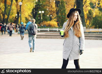 Sunny lifestyle portrait of young stylish hipster woman walking on street, wearing cute trendy hat, drinking hot latte coffee outdoors. Fashion blogger outfit. Photo toned style instagram filters