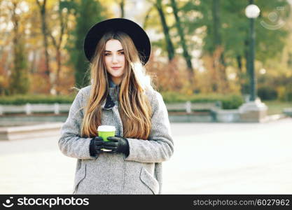 Sunny lifestyle portrait of young stylish hipster woman walking on street, wearing cute trendy hat, drinking hot latte coffee outdoors. Fashion blogger outfit. Photo toned style instagram filters