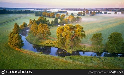 Sunny summer calm morning panorama. Small river, fog sunrise aerial view. First sunrays over green meadow and field