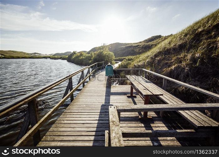 Sunny view of tourist sightseeing path along lakeshore. Rest place on wooden walkway in national reserve. Picturesque view of preserved landscape in evening.