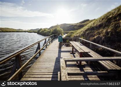 Sunny view of tourist sightseeing path along lakeshore. Rest place on wooden walkway in national reserve. Picturesque view of preserved landscape in evening.