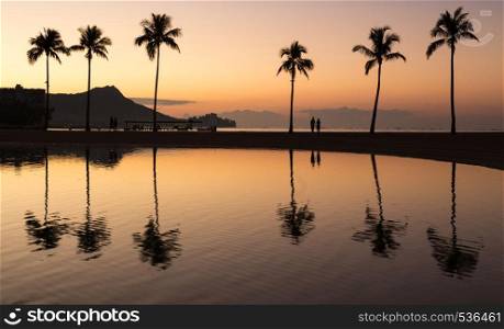 Sunrise in Waikiki with Diamond Head reflected in calm waters of pond as walkers walk along the beach. Sunrise over ocean with palm trees in Waikiki Hawaii