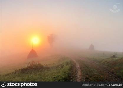 Sunrise over mountain field. Haystacks and road in misty rural hills. Foggy autumn morning in mountains.