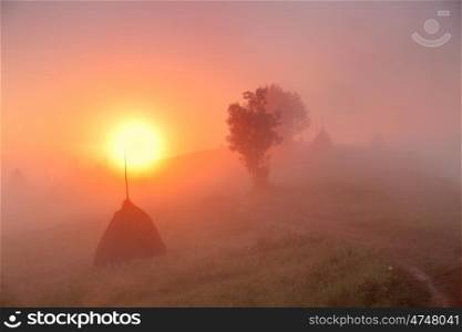 Sunrise over mountain field. Haystacks in misty rural hills. Foggy autumn morning in mountains.