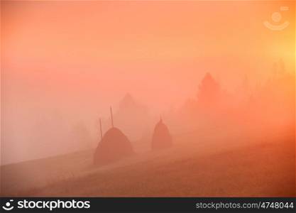 Sunrise over mountain field. Haystacks in misty rural hills. Foggy autumn morning in mountains.