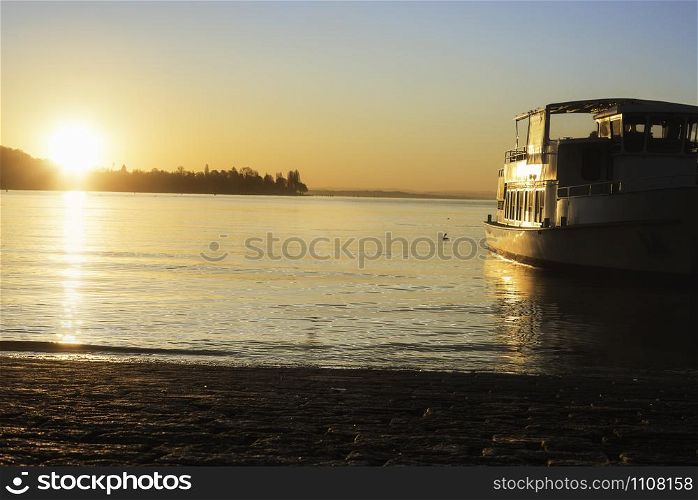 Sunrise over water and a boat on Constance lake, in Konstanz, Germany. Bodensee lakeshore and a ship cruising towards the sunrise. Water navigation.
