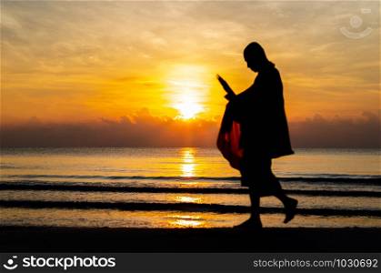 Sunrise with reflection on the sea and beach that have blurred silhouette photo of buddhist monk walking alms offering food in the morning on beach of Thailand.