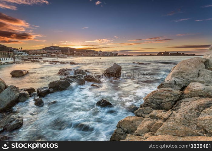 Sunset and clear skies over the town of Ile Rousse on the west coast of the Balagne region of Corsica with rocks and mediterranean sea in foreground and lights coming on in the town.