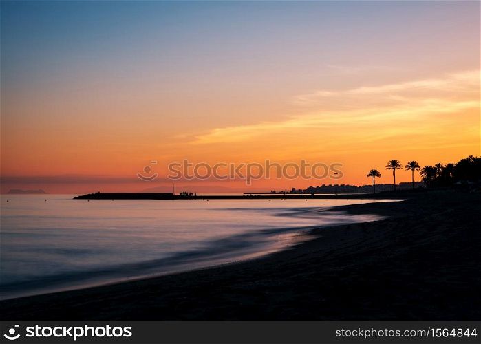Sunset at a beach in the Costa del Sol in Marbella, Spain, with the silhouette of a pier and Gibraltar to be recognised in the background. Long exposure.