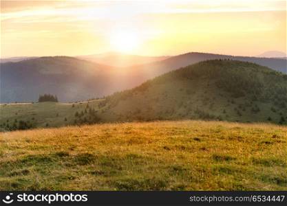 Sunset in mountains with forest, green grass and big shining sun on dramatic sky. Sunset in the mountains