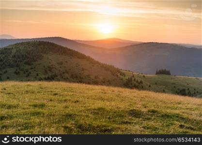 Sunset in the mountains with forest, green grass and big shining sun on dramatic sky. Sunset in the mountains