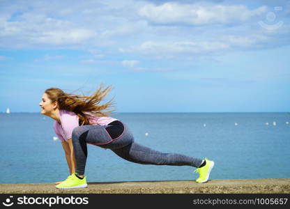 Super fit attractive young woman wearing fashionable outfit working out being active outside during sunny weather. Stretching her legs or practice yoga next to sea. Woman stretching legs next to sea