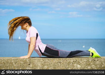 Super fit attractive young woman wearing fashionable outfit working out being active outside during sunny weather. Stretching her back or practice yoga next to sea. Woman doing yoga next to sea