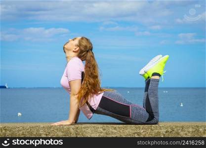 Super fit attractive young woman wearing fashionable outfit working out being active outside during sunny weather. Stretching her back or practice yoga next to sea. Woman doing yoga next to sea