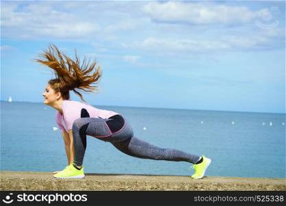 Super fit attractive young woman wearing fashionable outfit working out being active outside during sunny weather. Stretching her legs or practice yoga next to sea. Woman stretching legs next to sea