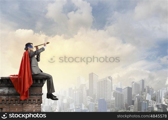Super hero sitting on top of building and looking in spyglass. Guy in super hero costume