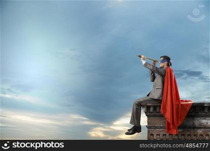 Super hero sitting on top of building and looking in spyglass. Guy in super hero costume