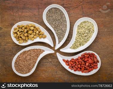 superfood samples (mulberry, chia seeds, hemp seeds, goji berry, flax seed) in teardrop shaped bowls against grunge wood