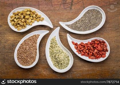 superfood samples (mulberry, chia seeds, hemp seeds, goji berry, flax seed) in teardrop shaped bowls against grunge wood