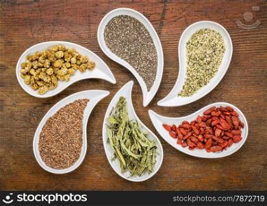 superfood samples (mulberry, chia seeds, hemp seeds, goji berry, stevia leaf, flax seed) in teardrop shaped bowls against grunge wood