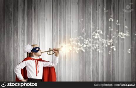 Superkid with trumpet. Cute girl of school age in superhero costume playing trumpet
