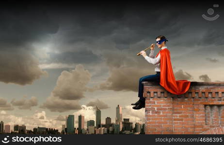 Superman looking in spyglass. Young man in superhero costume on top of building