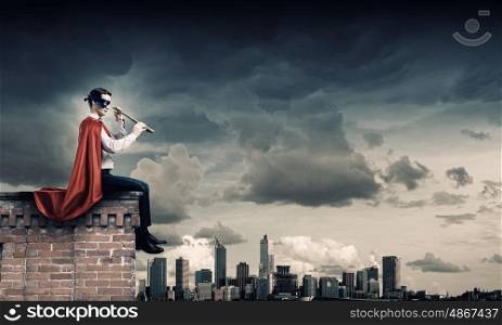Superman looking in spyglass. Young man in superhero costume on top of building