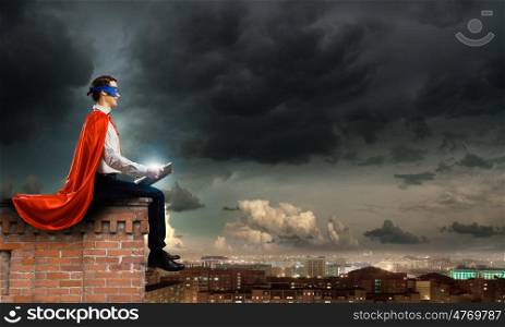 Superman with book. Superman in cape and mask sitting on top of building and reading book