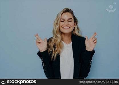 Superstitious young student girl with blond hair crossing fingers with both hands, invoking good luck before important exam or event, hopes for victory and success, feeling hopeful closing her eyes. Superstitious student girl with blond hair crossing fingers with both hands, invoking good luck